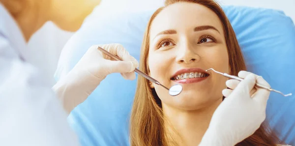 Güneşli diş kliniğinde dişçi tarafından incelenen diş telleriyle gülümseyen genç bir kadın. Sağlıklı diş ve ilaç konsepti — Stok fotoğraf