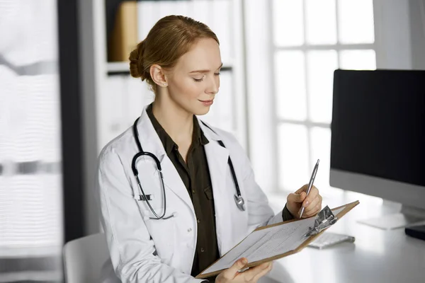 Alegre sonriente doctora usando portapapeles en la clínica. Retrato de mujer médica amigable en el lugar de trabajo. Servicio médico perfecto en el hospital. Concepto de medicina — Foto de Stock