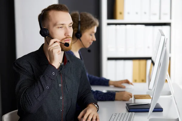 Homme d'affaires à barbe rouge parlant par casque près de sa collègue féminine alors qu'il était assis dans un bureau moderne. Groupe de personnes diversifiées au centre d'appels. Télémarketing et service à la clientèle — Photo