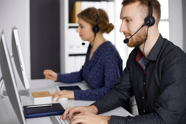 Homme d'affaires à barbe rouge parlant par casque près de sa collègue féminine alors qu'il était assis dans un bureau moderne. Groupe de personnes diversifiées au centre d'appels. Télémarketing et service à la clientèle — Photo