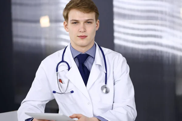 Blond mężczyzna lekarz siedzi i pracuje z tabletem w klinice w jego miejscu pracy, zbliżenie. Młody lekarz w pracy. Doskonała obsługa medyczna, koncepcja medycyny — Zdjęcie stockowe