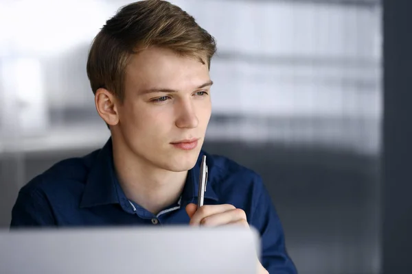 Jeune homme d'affaires blond réfléchissant à la stratégie à son lieu de travail avec ordinateur. Démarrage d'entreprise signifie travailler dur et hors du temps pour la réussite — Photo