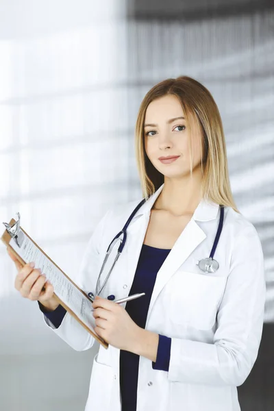 若いインテリジェントな女性医師は、診療所のキャビネットに立っている間、クリップボードを使用して、患者のためにいくつかの薬を処方しています。女性医師とともに聴診器で彼女の職場で — ストック写真