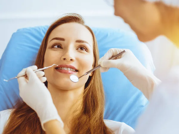 Glimlachende jonge vrouw met orthodontische haakjes onderzocht door tandarts in zonnige tandheelkundige kliniek. Gezonde tanden en geneeskundeconcept — Stockfoto