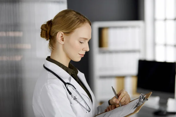 Friendly smiling female doctor using clipboard in clinic. Portrait of friendly physician woman at work. Medical service in hospital. Medicine concept