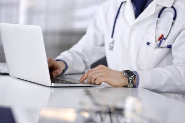 Doktor klinikte dizüstü bilgisayarla oturup çalışıyor, yakın plan. İşteki genç doktor. Mükemmel sağlık hizmeti, tıp konsepti