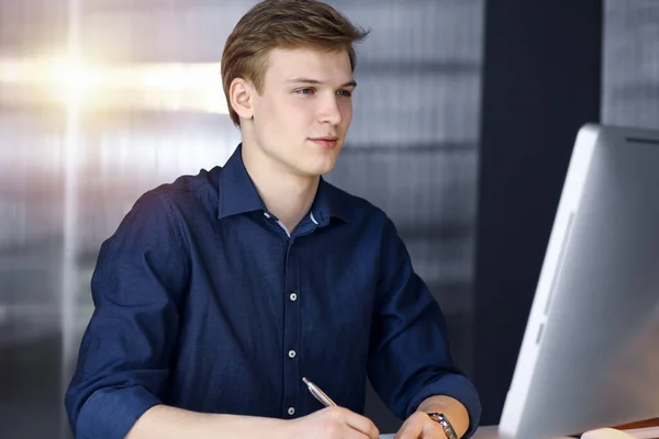 Jeune homme d'affaires blond réfléchissant à la stratégie sur son lieu de travail avec ordinateur dans un bureau sombre, éblouissement de la lumière sur le fond — Photo