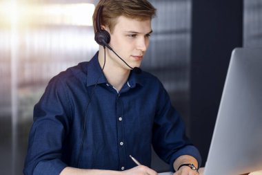 Genç sarışın işadamı, karanlık bir ofiste kulaklık ve bilgisayar kullanıyor, arka planda ışık parlıyor. Girişim işi çok çalışmak demektir.