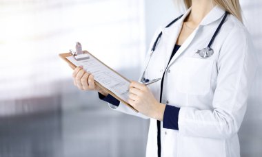 Bilinmeyen kadın-doktor klinikteki güneşli dolapta dururken bir not panosu kullanarak bazı notlar yazıyor. Steteskoplu bir kadın doktor, yakın plan. Hastanede mükemmel bir tıbbi hizmet.