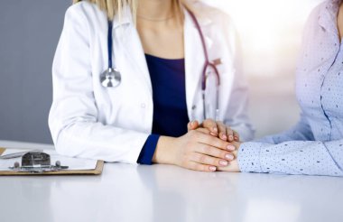 Bilinmeyen kadın doktor, bir klinikteki güneşli dolapta yan yana otururken hastasına güven vermek için elini tutuyor. Kadın hekim bir pano ve steteskop kullanıyor.