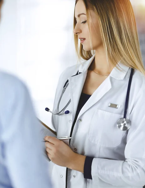 La jeune femme-médecin et sa patiente discutent de l'examen de santé actuel des patients, tout en restant ensemble dans un bureau d'hôpital ensoleillé. Médecin féminin écrit quelques marques, en utilisant un presse-papiers — Photo