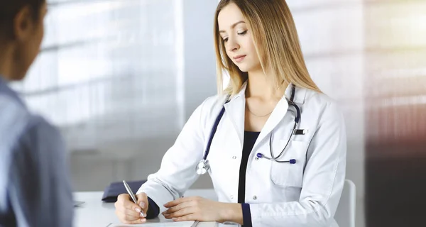 Unbekannte Ärztin schreibt ihrem Patienten ein paar medizinische Empfehlungen, während sie zusammen am Schreibtisch im sonnigen Schrank einer Klinik sitzen. Arzt benutzt Klemmbrett, Nahaufnahme — Stockfoto