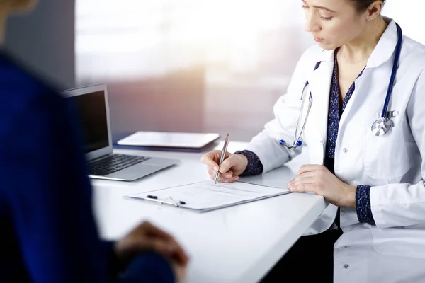 Donna-medico sconosciuto sta scrivendo alcune raccomandazioni mediche per la sua paziente, mentre sono seduti insieme alla scrivania nel gabinetto soleggiato in una clinica. Il medico sta usando un blocco appunti, primo piano — Foto Stock