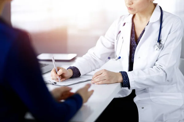 Unknown woman-doctor is writing some medical recommendations to her patient, while they are sitting together at the desk in the sunny cabinet in a clinic. Physician is using a clipboard, close-up — Stock Photo, Image