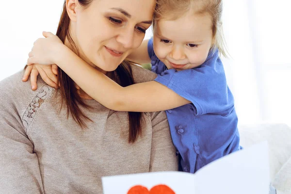Mothers day concept. Child daughter congratulates mom and gives her postcard with red heart shape. Mum and girl happy smiling and hugging. Family fun and holiday — Stock Photo, Image