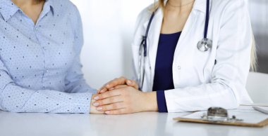 Bilinmeyen kadın doktor, bir klinikteki dolapta yan yana otururken hastasına güven vermek için elini tutuyor. Kadın doktor bir dosya ve steteskop kullanıyor. Kapat.