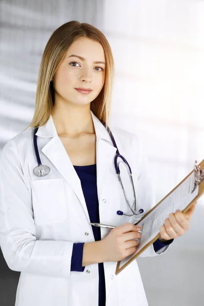 Una joven doctora optimista sostiene un portapapeles en sus manos, mientras está parada en un gabinete soleado de una clínica. Retrato de una médica amigable con un estetoscopio. Servicio médico perfecto en un — Foto de Stock