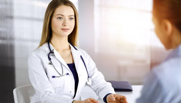 Jeune belle femme-médecin et sa patiente discutent de l'examen de santé actuel des patients, tout en étant assis ensemble au bureau dans l'armoire ensoleillée dans une clinique. Médecin féminin écrit certains — Photo