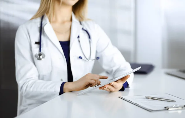 Jovem médica desconhecida está verificando alguns nomes de medicamentos, enquanto se senta na mesa em seu gabinete em uma clínica. O médico com um estetoscópio está usando um computador tablet, close-up. Perfeito médico — Fotografia de Stock