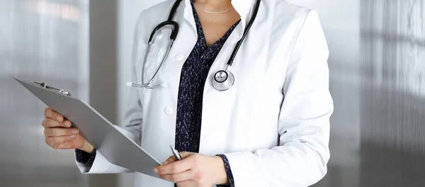 Χαρούμενη έξυπνη γυναίκα-γιατρός κρατά ένα πρόχειρο στα χέρια της, ενώ στέκεται σε ένα γραφείο κλινική. Πορτρέτο της φιλικής γιατρού γυναίκα με στηθοσκόπιο. Τέλεια ιατρική εξυπηρέτηση σε νοσοκομείο — Φωτογραφία Αρχείου