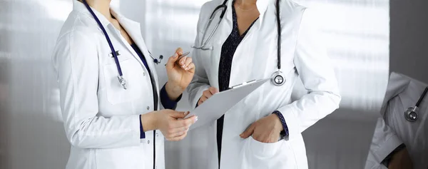 Two female physicians are discussing their patients medical tests, while standing in a clinic office. Doctors use a clipboard at work. Teamwork in medicine