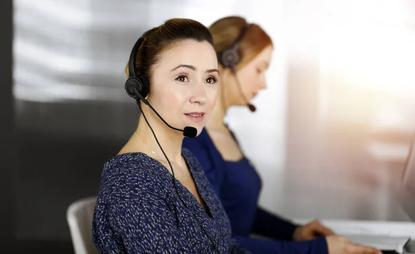 Two busineswomen have conversations with the clients by headsets, while sitting at the desk in a sunny modern office. Diverse people group in a call center. Telemarketing and customer service