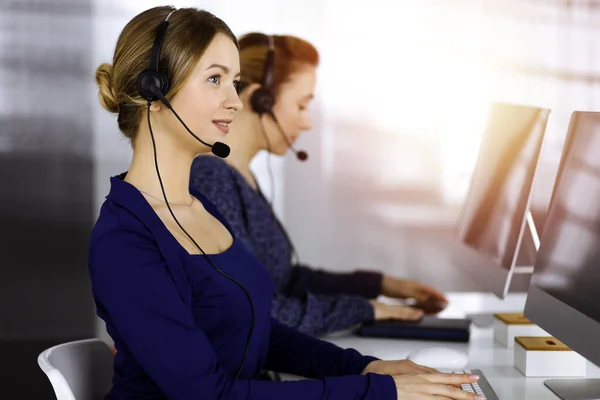 Deux femmes d'affaires ont des conversations avec les clients par écouteurs, tout en étant assis au bureau dans un bureau moderne ensoleillé. Groupe de personnes diverses dans un centre d'appels. Télémarketing et service à la clientèle — Photo