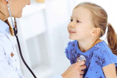 Doktor steteskopla küçük bir kızı muayene ediyor. Her zamanki tıbbi muayenede gülümseyen çocuk hasta. Tıp ve sağlık hizmetleri kavramları