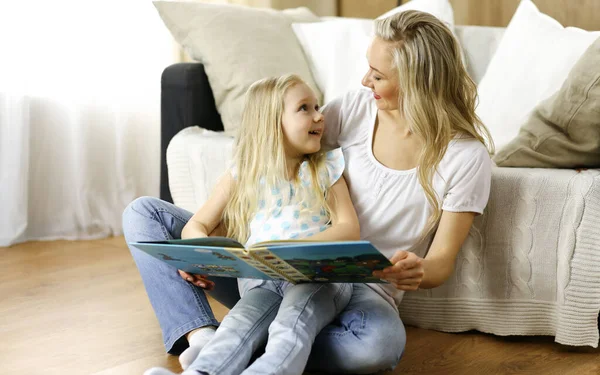 Happy family. Blonde young mother reading a book to her cute daughter while sitting at wooden floor. Motherhood concept