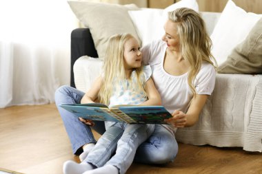 Mutlu bir aile. Sarışın genç anne ahşap zeminde otururken sevimli kızına kitap okuyor. Annelik kavramı