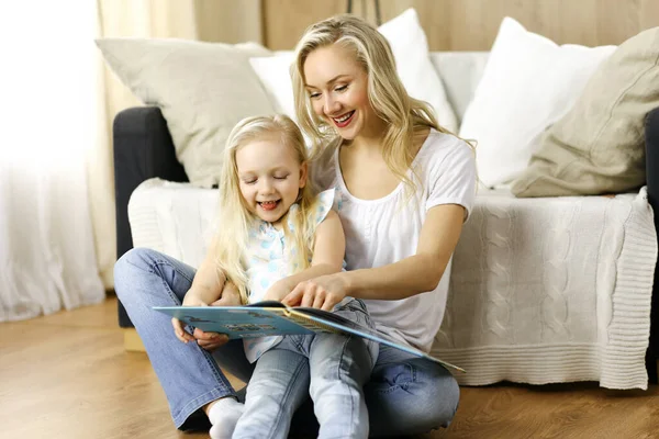 Bonne famille. Jeune mère blonde lisant un livre à sa jolie fille assise au sol. Concept de maternité — Photo