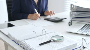 Ofis çalışanı, iş yerinde dizüstü bilgisayarla, büyüteçle ve belgelerle, dosyalarla ve gözlüklerle notlar hesaplıyor ve yazıyor. Denetim ve vergi kavramı