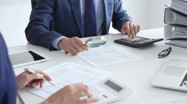 Erkek finansal analistler, iş yerinde bir iş arkadaşıyla kurumsal verileri incelerken büyüteç ve hesap makinesi kullanarak belgeleri inceliyorlar. Denetim ve vergi kavramı.