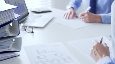 Ofis çalışanları mali raporları analiz ediyor ve ön planda evrak dolu mavi dosyalarla dolu beyaz bir masada otururken belgeleri imzalıyorlar. İş dünyasında denetim ve vergi teması