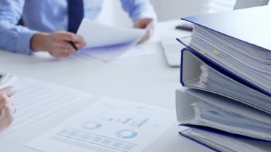 Ofis çalışanları mali raporları analiz ediyor ve ön planda evrak dolu mavi dosyalarla dolu beyaz bir masada otururken belgeleri imzalıyorlar. İş dünyasında denetim ve vergi teması