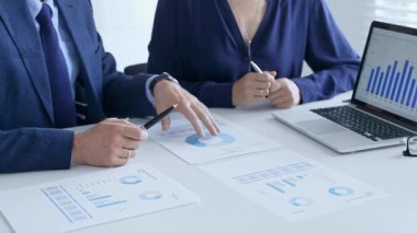 Bir proje üzerinde işbirliği yapan iş arkadaşları, dizüstü bilgisayar ve belgelerdeki mali verileri analiz ederken çizelgelere işaret ediyorlar. İş adamları ve finans kavramı