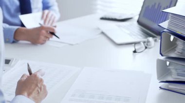 Ofis çalışanları mali raporları analiz ediyor ve ön planda evrak dolu mavi dosyalarla dolu beyaz bir masada otururken belgeleri imzalıyorlar. İş dünyasında denetim ve vergi teması