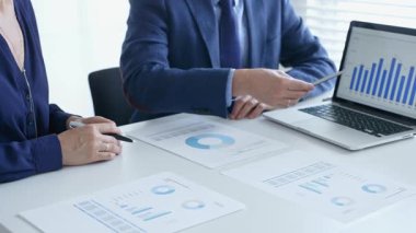 Bir proje üzerinde işbirliği yapan iş arkadaşları, bir dizüstü bilgisayar ve belgelerde yer alan mali tablolar ve grafikleri analiz ederken, bir toplantı sırasında önemli verilere işaret ediyorlar. İş adamları ve finans kavramı