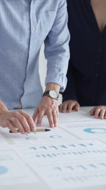 İş arkadaşları mali raporları inceliyor ve masadaki çizelge ve grafiklere işaret ediyor. Verileri analiz etmek ve stratejileri planlamak için birlikte çalışıyorlar.