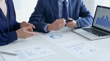 Bir proje üzerinde işbirliği yapan iş arkadaşları, bir dizüstü bilgisayar ve belgelerde yer alan mali tablolar ve grafikleri analiz ederken, bir toplantı sırasında önemli verilere işaret ediyorlar. İş adamları ve finans kavramı