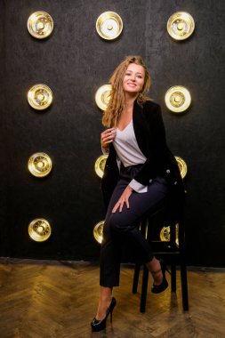 Siyah ceketli ve topuklu ayakkabılı zarif sarışın bayan. Moda stüdyosunda moda kadını. Arkaplanda yaratıcı lambalar.