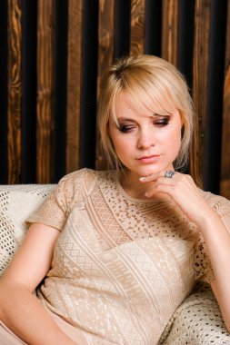 Klasik bej elbiseli zarif bir bayan. Kaküllü, büyük deri sandalyede oturan şık bir kadın. Arkaplanda tahta duvar var..