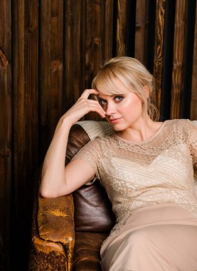 Klasik bej elbiseli zarif bir bayan. Kaküllü, deri sandalyede oturan şık bir kadın. Arkaplanda tahta duvar var..