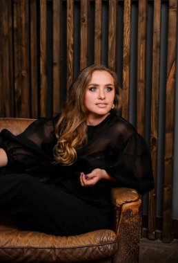 Siyah bluzla poz veren zarif sarışın bayan. Büyük deri sandalyede oturan şık bir kadın. Arkaplanda tahta duvar var..