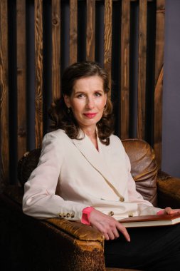 Zarif, beyaz takım elbiseli, defterli bir iş kadını. Psikoterapist yaşam tarzı. Büyük deri sandalyede oturan kadın. Arkaplanda tahta duvar var..