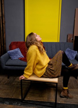 Sarı kazaklı, etekli ve kahverengi çizmeli bir kızın portresi. İçerideki masada oturan kadın. Arkaplanda sarı kare.