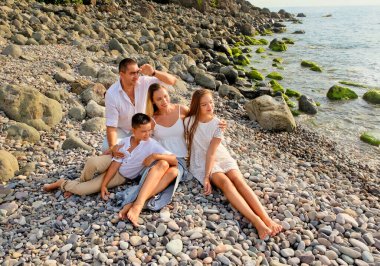 Neşeli, mutlu aile, baba, anne, oğul ve kız taş deniz sahilinde dinleniyor. Beyaz elbiseli bir aile kıyı şeridinde eğleniyor. Yaz günü ve tatil zamanı konsepti.