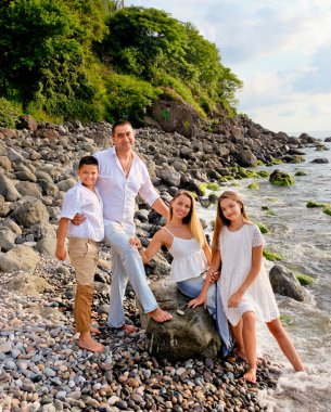 Neşeli, mutlu aile, baba, anne, oğul ve kız taş deniz sahilinde dinleniyor. Beyaz elbiseli bir aile kıyı şeridinde eğleniyor. Yaz günü ve tatil zamanı konsepti.