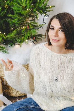 Mutlu, çekici bir kadının portresi. Evde yerde Noel ışığı var. Arka planda minimalist Noel ağacı var. Kabarık tüylü, beyaz kazaklı esmer kız. Yeni yıl arifesi havası..