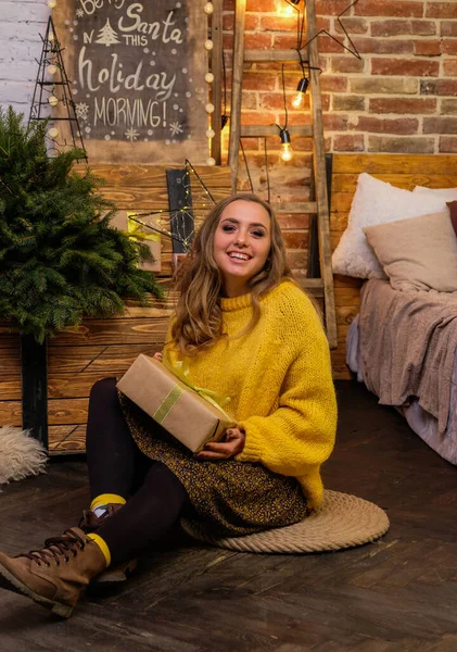 Portrait of happy, attractive woman with christmas gift on floor at home. Loft style christmas cozy decor on the background. Blonde, cute girl in fluffy, yellow oversized sweater. New Year\'s eve mood.